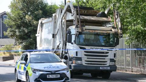 A white police car parked next to a white bin lorry in a street with blue and white police tape in front of them