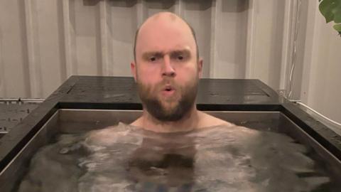 A man breaths out whilst sitting in a cold water bath.