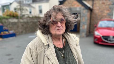 Corrine Joy wearing a dark top and a cream coloured coat. She has dark brown/grey hair.