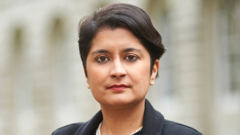 Shami Chakrabarti is looking at the camera and has short brown hair and brown eyes, and is wearing a black coat. The background behind her is blurred.