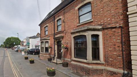 Brick buildings with bay windows and a step with railings leading to the front door, which is painted black, and there are flowers in pots on the pavement and hanging baskets by the door. The building is on a road with double yellow lines and there are newer buildings in the background, with a tree.