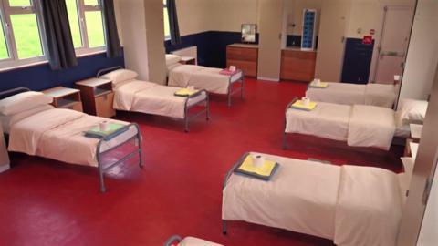 A dormitory-style room with multiple single beds is neatly made and spaced apart on a red floor, each bed with bedding and personal items on a tray, inside a former military building.