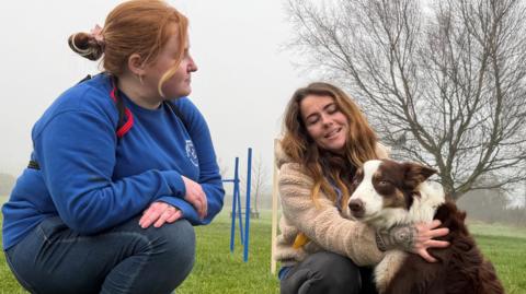 A woman with red hair and a blue jumper crouches next to another woman with red hair and a beige jumper. Woman in beige jumper holds a brown border collie.