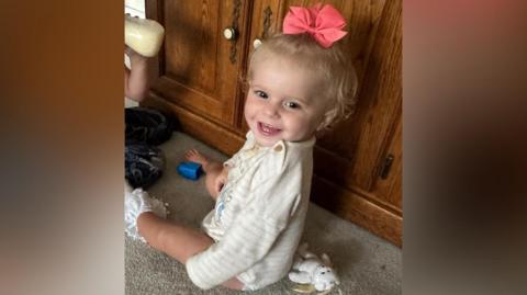 Viviana-Ray Butnaru - a young girl sitting on the floor, in front of a brown sideboard. She has turned her head to the left to smile at the camera. She has blonde hair with a pink bow in it and is wearing a white striped top.