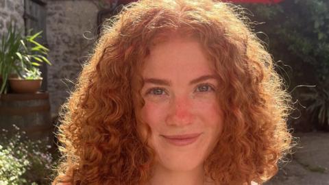 Gemma is a young woman, smiling into the camera. The sun is shining on her curly red hair. The background behind her is a stone wall with some green plants.