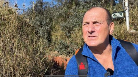 Toni Schofield squinting into the camera, wearing a blue fleece and a backpack, standing in front of a hedge and a public footpath sign