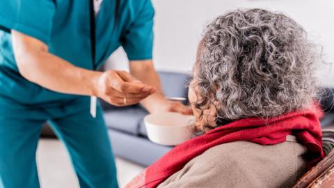 A standing, uniformed care worker feeds a seated, elderly woman from a bowl with a spoon