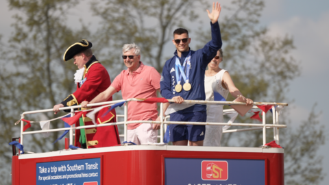 A man with dark glasses and dark hair, wearing two gold meda;s around his neck waves from a red podium.