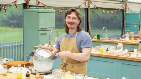 Iain working on a cake in the Bake Off tent. Iain has a blue top on and beige apron, long brown hair and a moustache. 