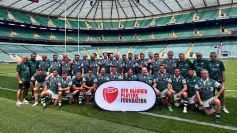 A rugby team is sitting on a rugby field in their kit. Some have their arms around each other. They are holding a sign with the RFU injured players foundation logo on it.