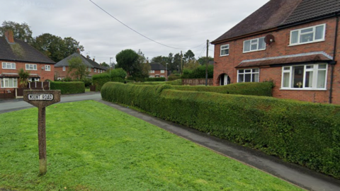 Semi-detached houses with bay windows sit behind neatly trimmed hedgerows.
A sign on a concrete post on a green in front of the houses says Mount Road.