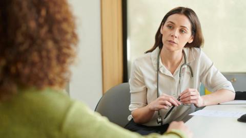 A GP wearing a stethoscope around her neck talks to a patient.