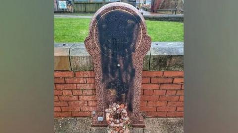 A memorial stone with black paint on it. The memorial has two wreaths at its foot, with a brick wall and green area behind it. 