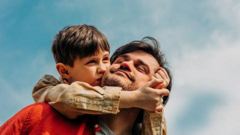 A father gives a piggy back to his son, the sky behind them is blue.