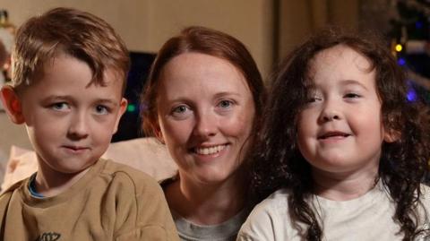 Youngsters Theo and Evie are sitting on their mother Chloe Green's lap and smiling brightly at the camera. There is a lit Christmas tree behind them.