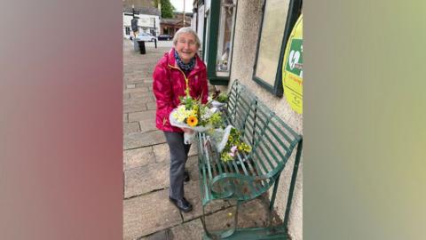 Anne Cradock is holding a bunch of flowers and is standing in front of a green metal bench which also has two bunches of flowers placed on it. Anne has short white/grey hair and is smiling broadly at the camera.