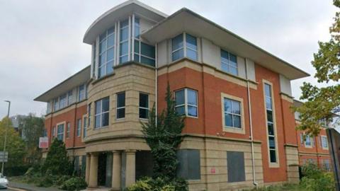 The front view of a large multi-storey building. There are trees and greenery surrounding it. 