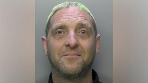 A mugshot shows an unshaven man with short bleached hair.