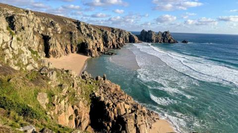 Overlooking Pedn Vounder beach. A rocky cliff face leads down to a sandy beach. Waves are rolling onto the shore. The sky is cloudy.