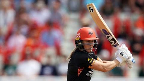 Ellyse Perry, wearing a black top and orange helmet, batting with her bat raised