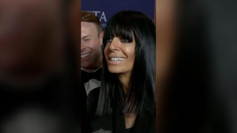 Claudia Winkleman smiling. She has a man stood behind her and a blue promotional Bafta backdrop.
