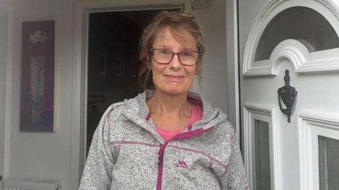A woman with brown hair tied up. She has glasses and is standing in a doorway with the white door open. She is wearing a grey and pink zip hoodie.