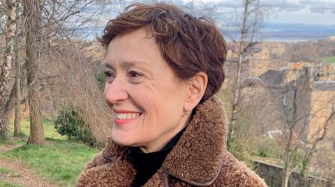 Carys Davies is smiling as she looks to the left of the camera. She has short brown hair and is wearing a black top with a brown fleece jacket. She is in the countryside, with trees behind her.