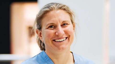 A woman with tied-back blonde hair, brown eyes and wearing a blue medical top. She is smiling at the camera with a blurred white background behind her.