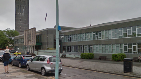 The image shows a street scene featuring an institutional or governmental building with multiple windows and a flagpole with a flag flying. There are cars parked along the street and people walking on the pavement. 