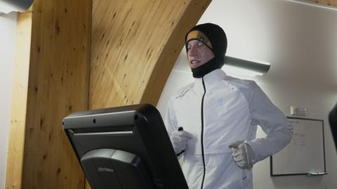 Logan running on a treadmill in multiple layers of clothing including white jacket plus yellow hat, black snood and white gloves to try to replicate the humid conditions of New Delhi