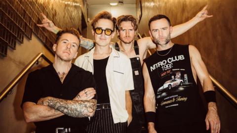A boy band of four people all stood in the middle of a stairway and pulling serious expressions. They all have short brown hair and the man on the left has his arms folded with tattoos, the man next to him is wearing yellow tinted glasses, the man next to him is wearing black dungarees and pulling a rock sign with both his hands, and the man next to him is wearing a black vest that has a car on it. The stairway is lit in a brown lighting.