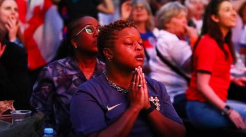 A woman in a blue England T-shirt holds her hands in the prayer position surrounded by fans in Boxpark, Croydon, London, in a particularly tense moment during the semi-final between England and Italy 