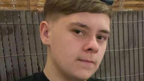A 15-year-old boy looks at the camera slightly sideways. He has a neutral expression. He wears a black t-shirt.