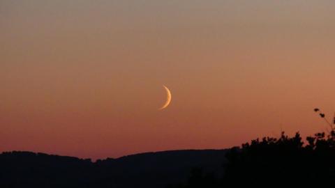As the sun sets, a crescent moon rises over the Somerset hills. The sky is still lit in red, orange and yellow hues from the sun, although the sun itself is not visible.