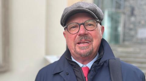 Deputy David Dorrity - A man with a check flat cap on and black rimmed glasses and a grey and black goatee beard. He is wearing a blue coat over a blue shirt with a red and white spotted tie. 