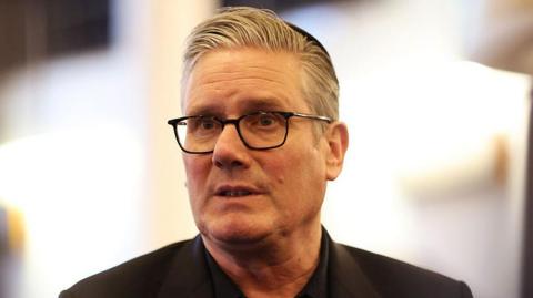 Headshot of Sir Keir Starmer, wearing a black jacket and shirt and looking slightly to his right, against a blurred background