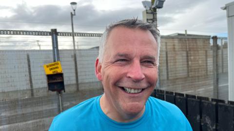 Gary Milling, governor of Magilligan Prison. He is smiling, has silver hair and wearing a blue t-shirt. Outside fencing of the prison can be seen in the background.