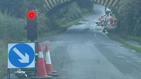 A temporary traffic light holding traffic on a road. In the background is the railway bridge with cars waiting on the other side.