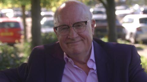 Sir Michael Lyons looking direct to camera, wearing a purple suit jacket and open collar pink shirt. He is smiling and wearing a pair of glasses. It is a sunny day and while there are trees behind him, there are also several cars parked in a surface car park, out of focus in the background