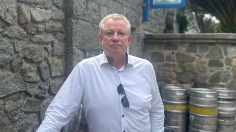 Tom Jones, a fair hared, freckled man in his 50s stood leaning against a bar cannister. He is wearing a white button shirt with black buttons and his sunglasses are hooked on his shirt. He is staring passed the lens. 