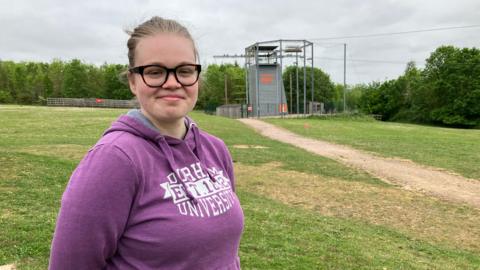 A woman with glasses in a purple hoodie looks at the camera. A tall climbing frame is in the background.