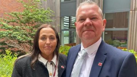 A woman with dark hair, a white blouse and dark jacket next to a man with a grey hair and a blue suit and white shirt in front of a building and some bushes