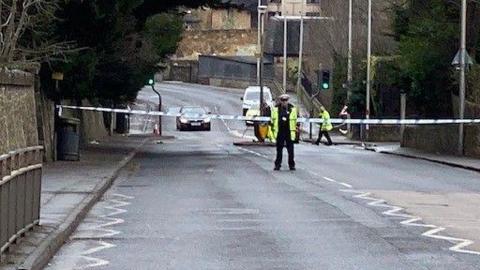 A police officer standing on a main road which is cordoned off with police tape. 