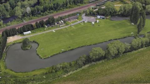 A view of Ferryboat Farm Fisheries. In the centre of the picture is an L-shaped pond surrounded by hedges and trees. A big lawn is occupied by a handful of caravans. A building and sheds can be seen just off the main road.