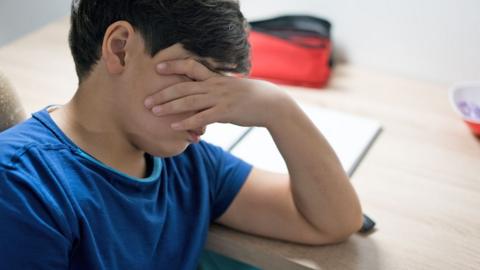 A child wearing a blue T-shirt sits at a wooden desk, covering his eyes with his left hand. He has brown hair and there is a notebook and pencil case on the desk in front of him.