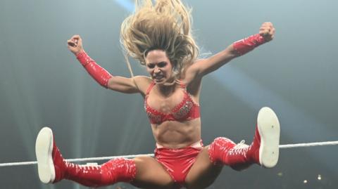 Maxxine Dupri is pictured in a really close-up shot, mid-jump in a wrestling ring. She is wearing a red bikini-type outfit with long, red socks and red trainers. She has long blonde hair which is pictured streaming above her head/