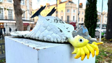 An artwork featuring a comic white cockatiel bird lying on its back on a stone plinth with a park and buildings in the background