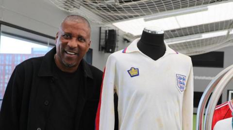 Viv Anderson, wearing a black jumper and coat, stands smiling beside the England shirt he wore on his debut in 1978. The V-neck jersey is white but slightly faded and has mud stains across the front. An England crest is on the left breast while an Admiral manufacturer's logo is on the opposite side. The long sleeves have red and blue stripes.