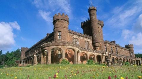 The sandstone castle has turrets and battlements. It is set among grassy grounds filled with flowers.
