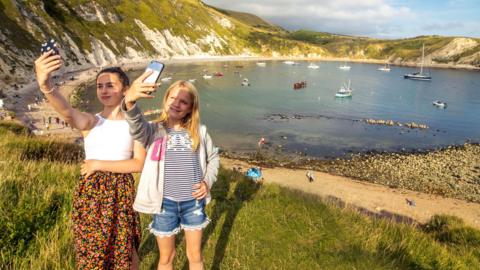 Two young girls hold up mobile phones on a grassy bank above Lulworth Cove, which is filled with small boats on a sunny day.